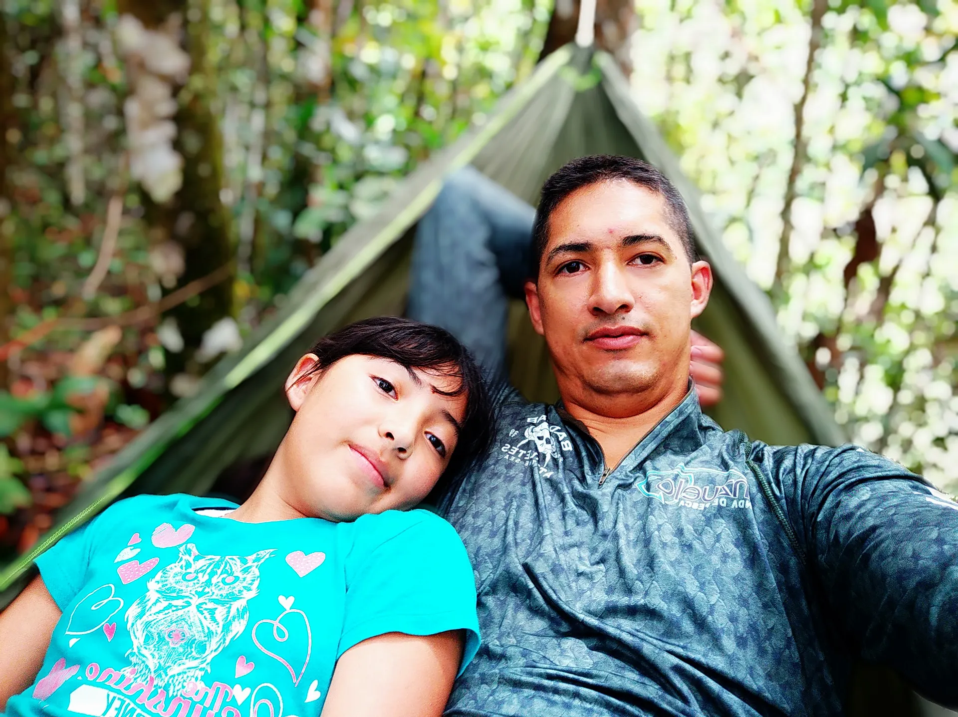 Un padre y su hija recostados juntos en una hamaca verde, en medio del bosque, mirando a la cámara.