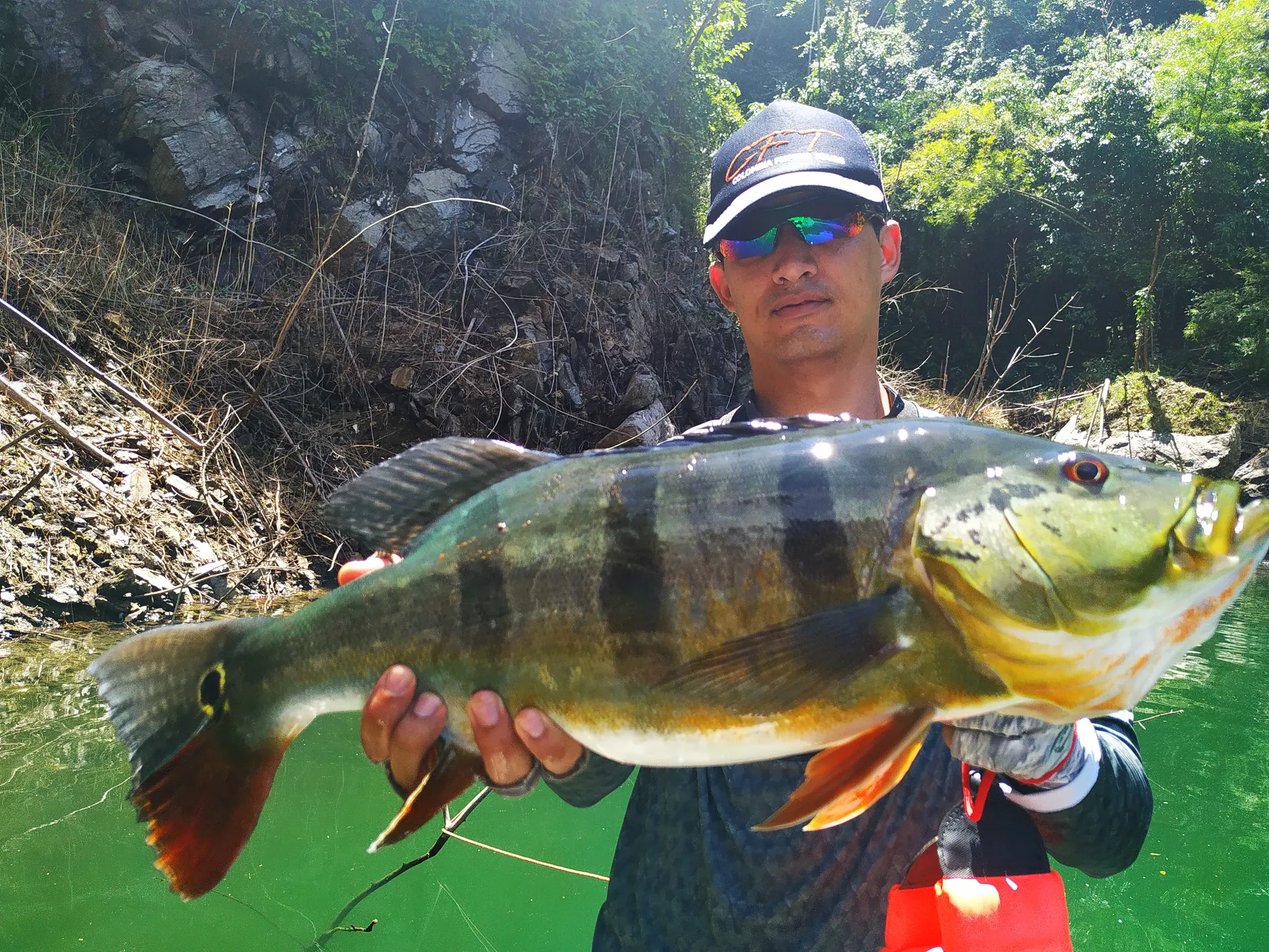 Un pescador con gorra sostiene con las dos manos un pavón grande de rayas oscuras y cola roja, junto a una pared rocosa a la orilla del embalse.