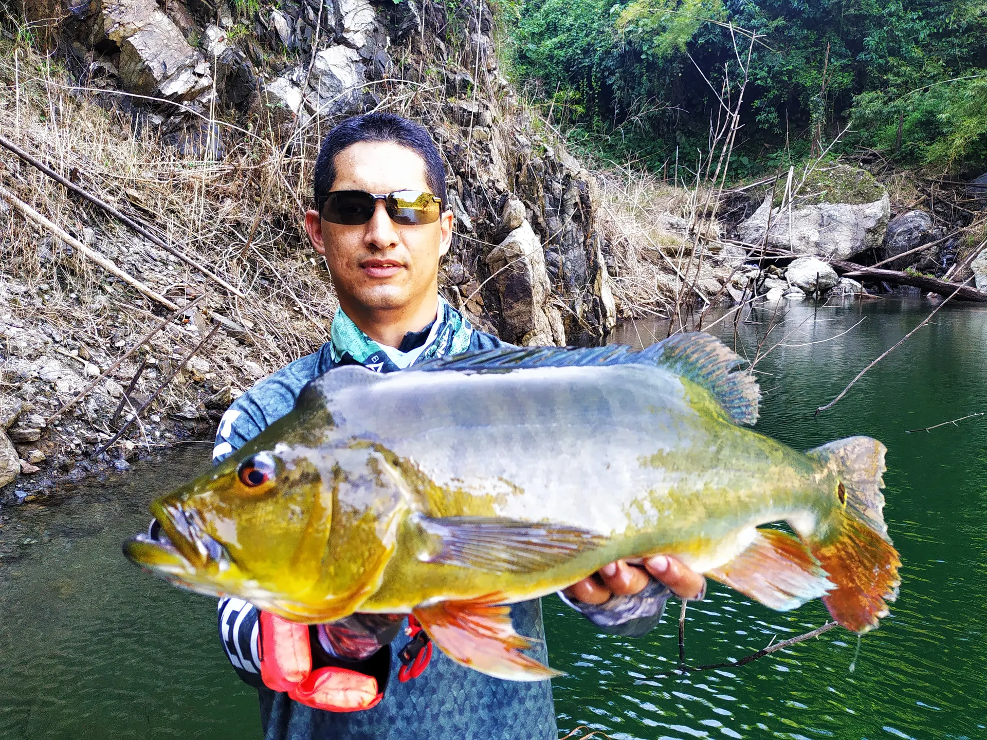 Un pescador con gafas de sol sostiene un pavón plateado y dorado con las dos manos, frente a una pared de roca junto al río.