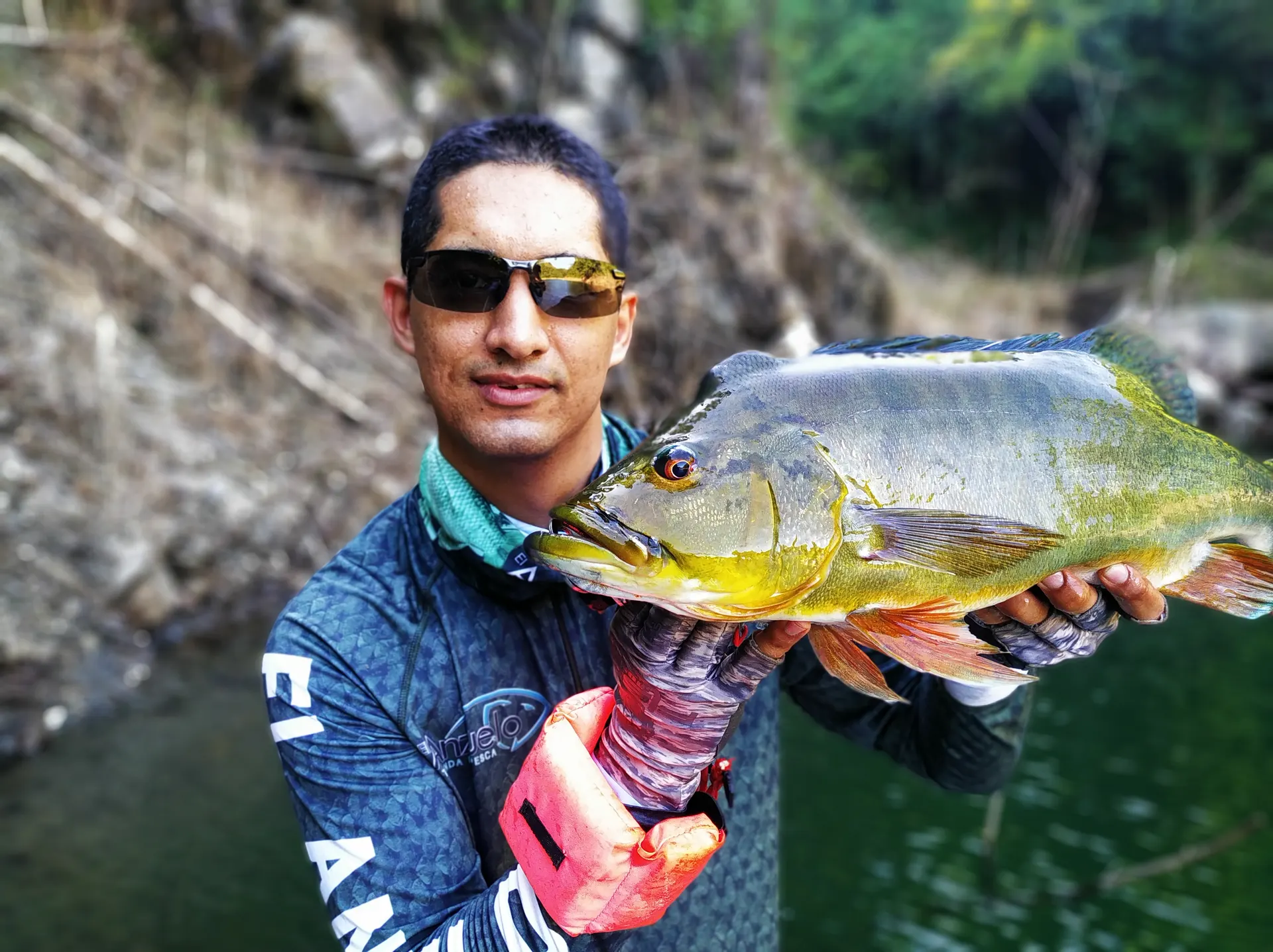 Un pescador con gafas de sol sostiene un pavón dorado con las dos manos, mirando a la cámara junto a la orilla rocosa.
