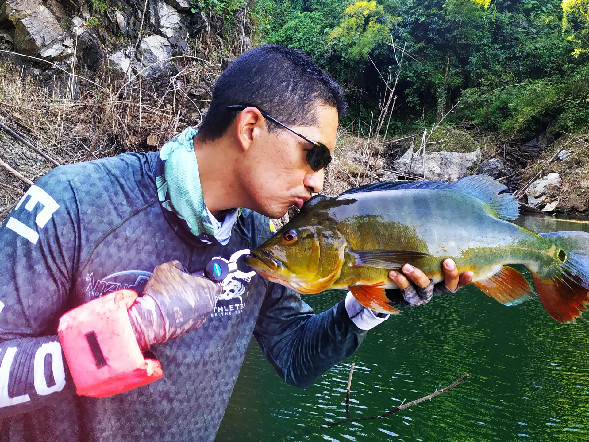 Un pescador le da un beso a un pavón dorado que sostiene con las dos manos, frente a una pared rocosa junto al río.