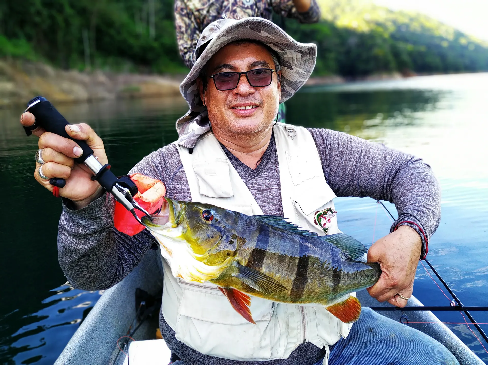 Un pescador con sombrero de ala ancha y gafas de sol sostiene un pavón de rayas oscuras, sentado en un bote sobre el embalse en calma.