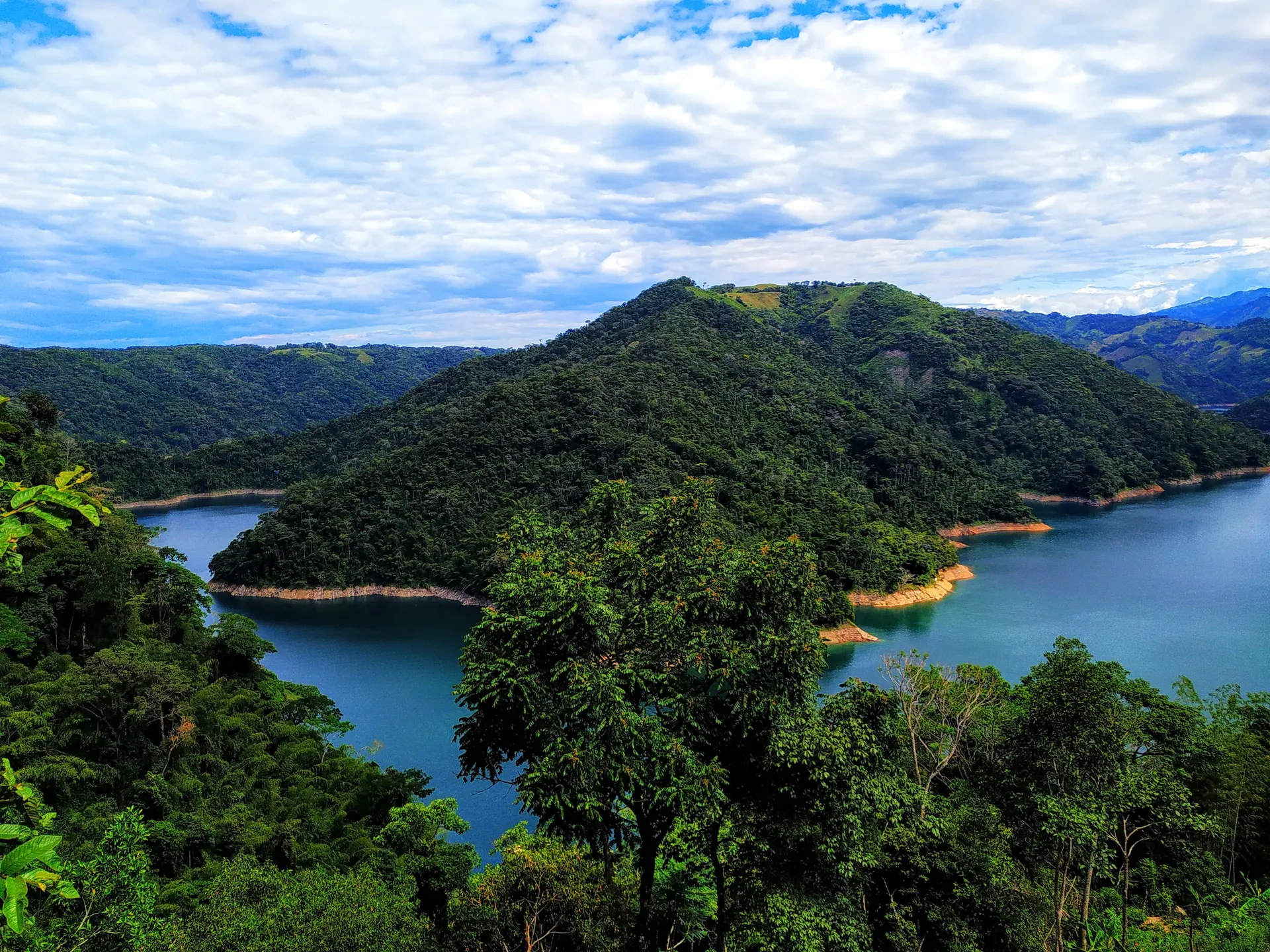 Vista aérea de un embalse azul serpenteando entre montañas cubiertas de selva, con la orilla anaranjada asomando bajo el nivel del agua.