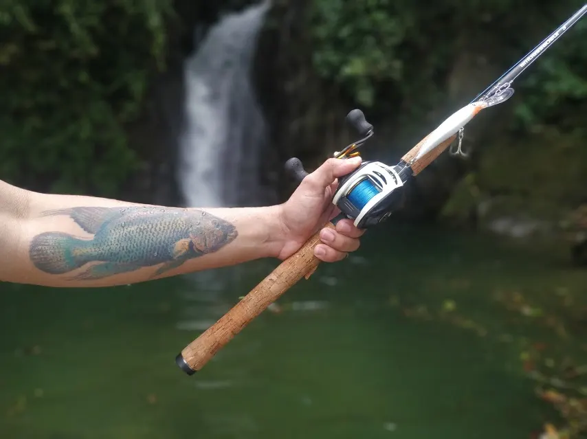 Un brazo con un tatuaje de pez sostiene una caña con carrete de perfil bajo, frente a una pequeña cascada en el bosque.