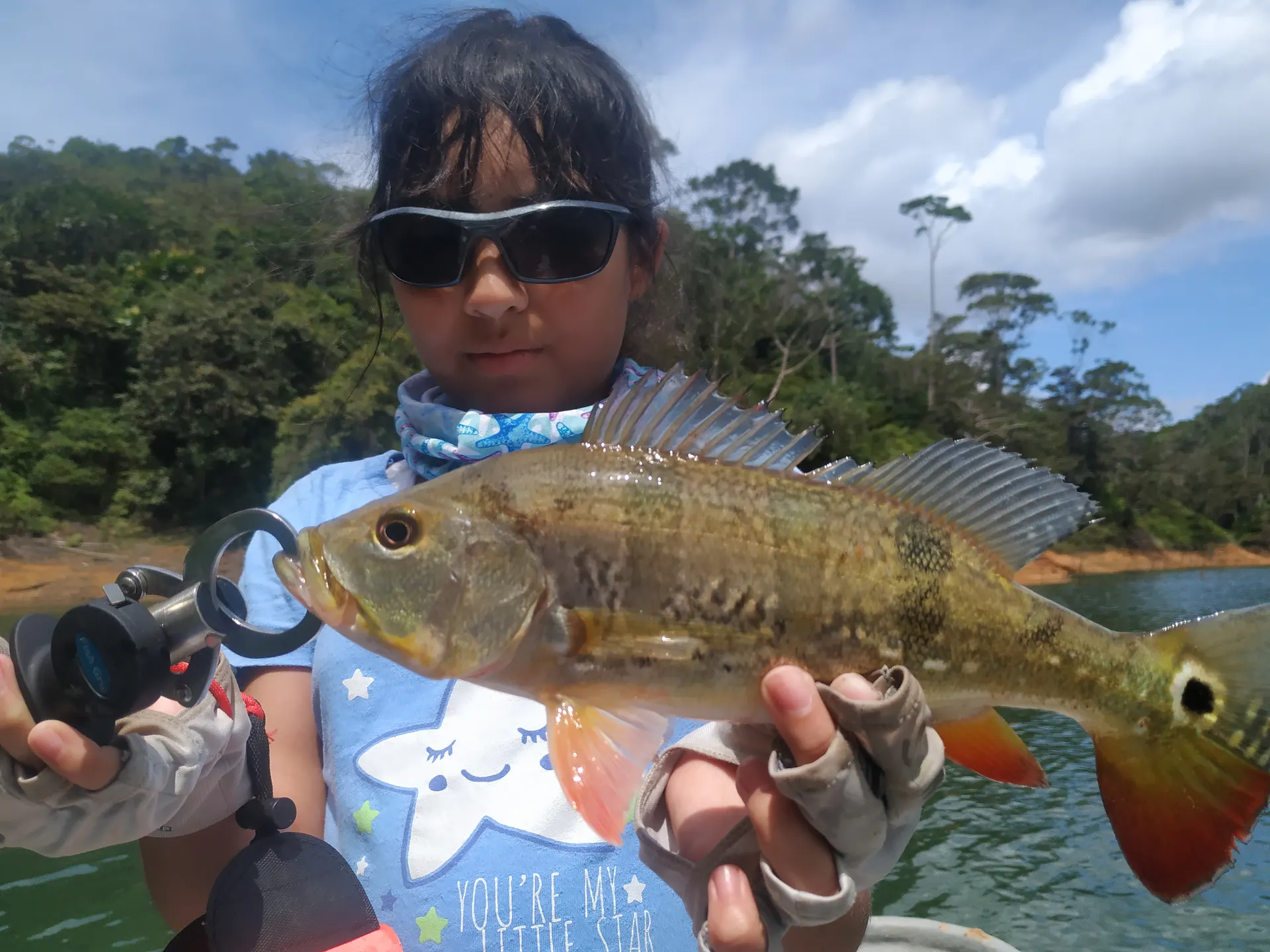 Una niña con gafas de sol sostiene con las dos manos su primer pavón, de aletas rojas y la mancha negra en la cola, en un embalse rodeado de selva.