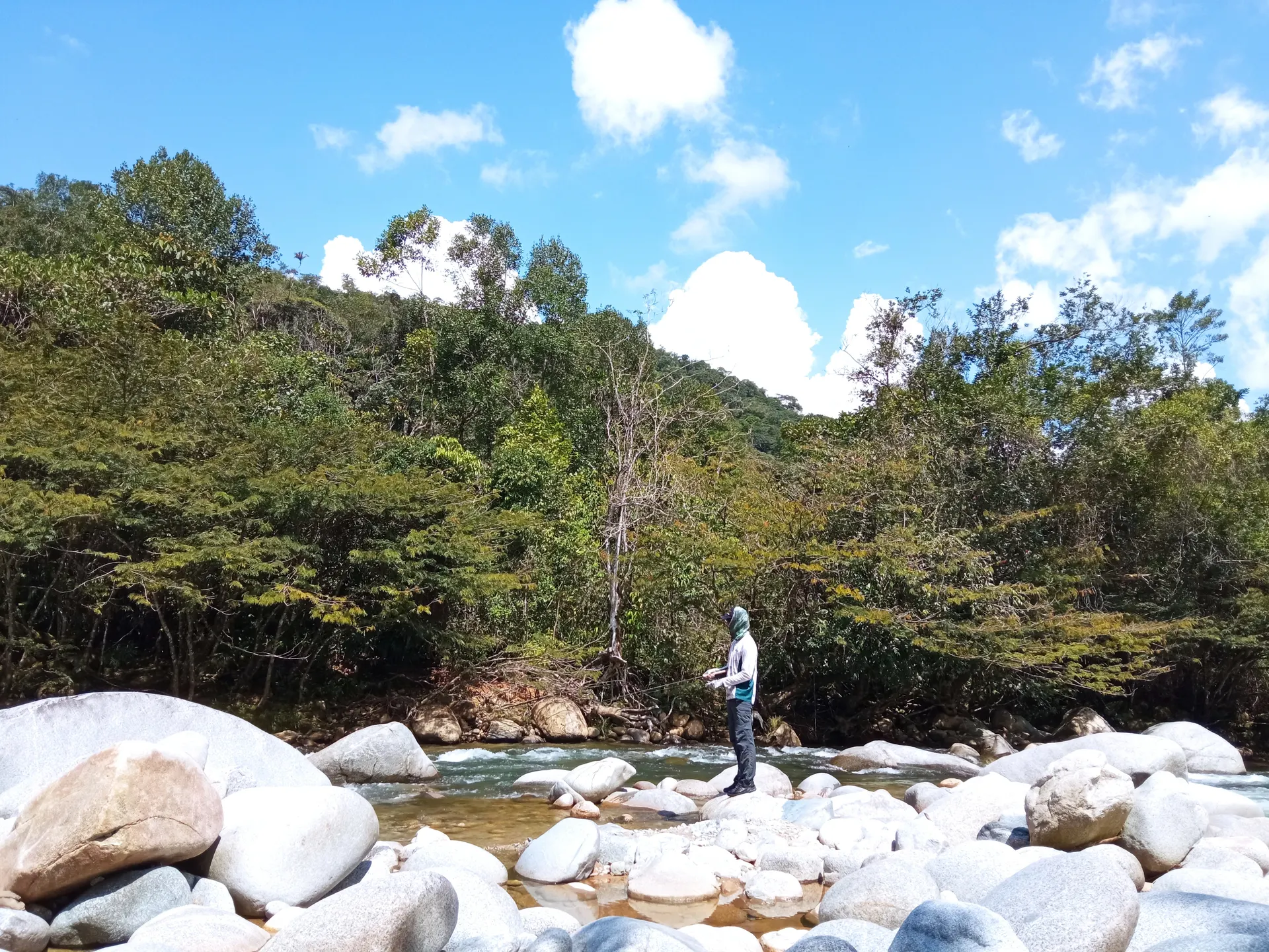 Un pescador con capucha lanza de pie sobre las piedras blancas de un río de montaña, rodeado de selva bajo un cielo azul con nubes.