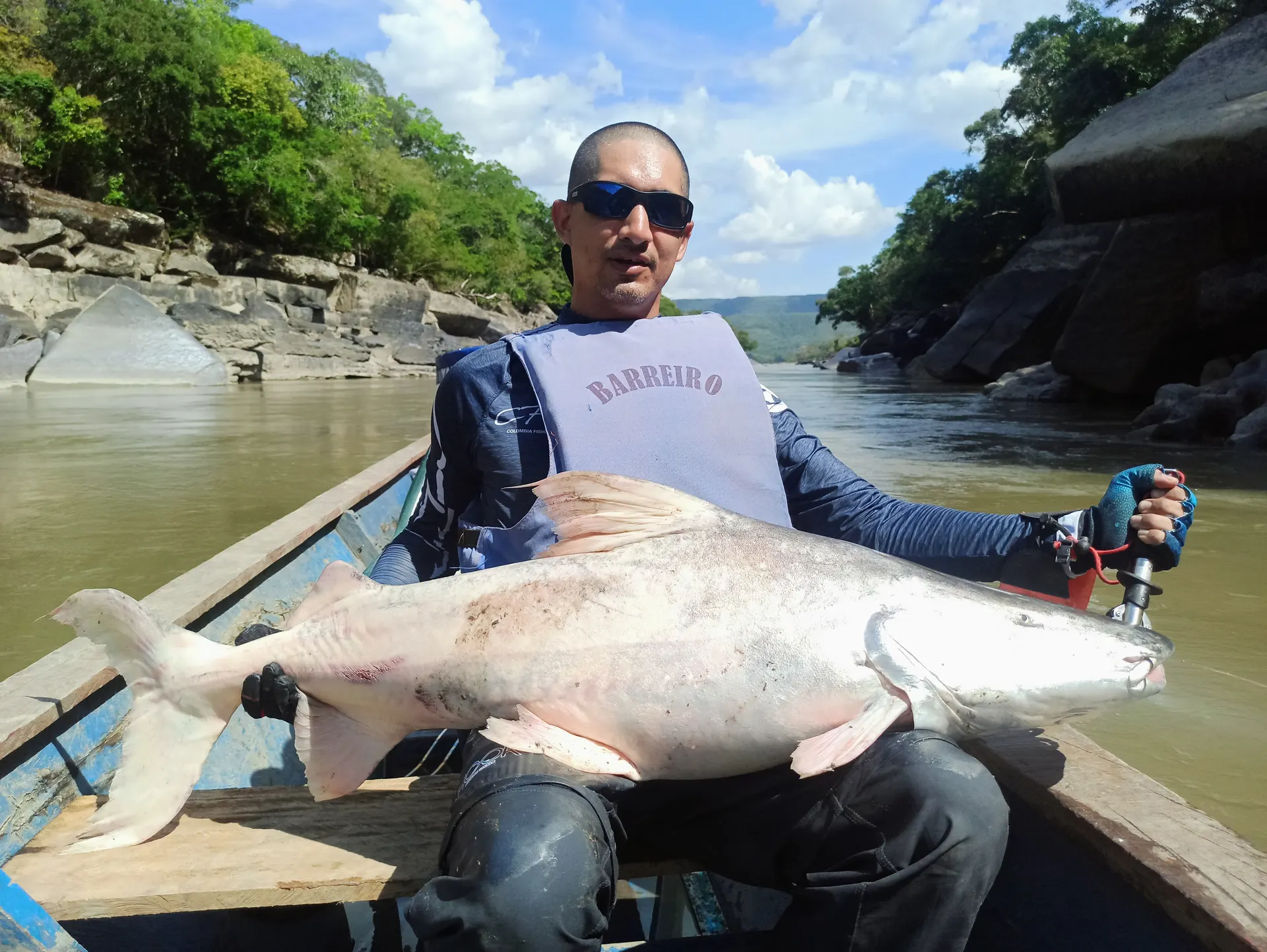Un pescador con chaleco salvavidas sostiene sobre sus piernas un bagre dorado enorme, sentado en una canoa en un río de aguas turbias entre farallones.