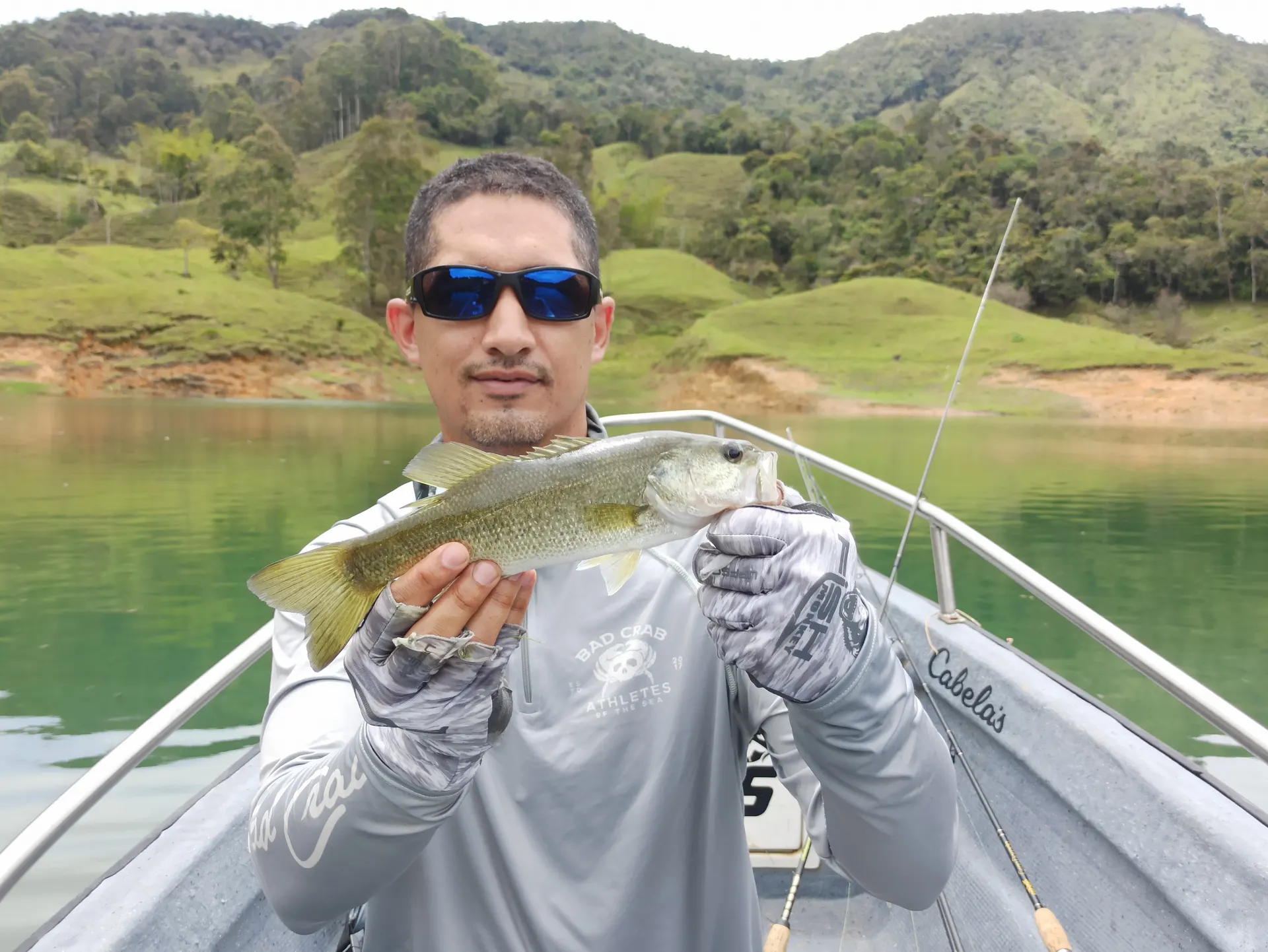 Un pescador con camiseta gris y gafas de sol sostiene un black bass desde la proa de un bote, con las colinas verdes del embalse al fondo.