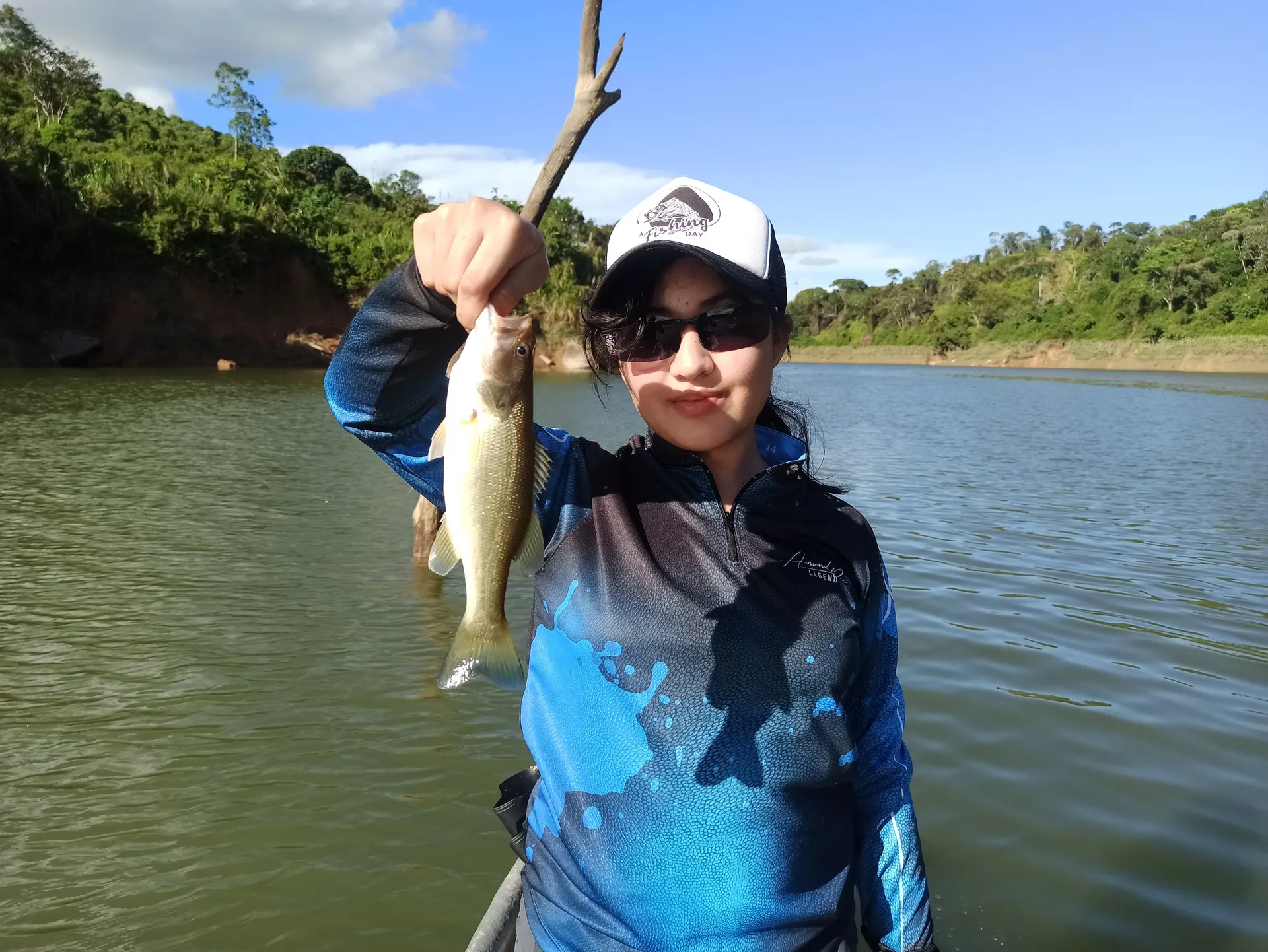 Una joven con gorra de A Fishing Day y gafas de sol levanta un black bass pequeño desde un bote, con la orilla selvática del embalse detrás.