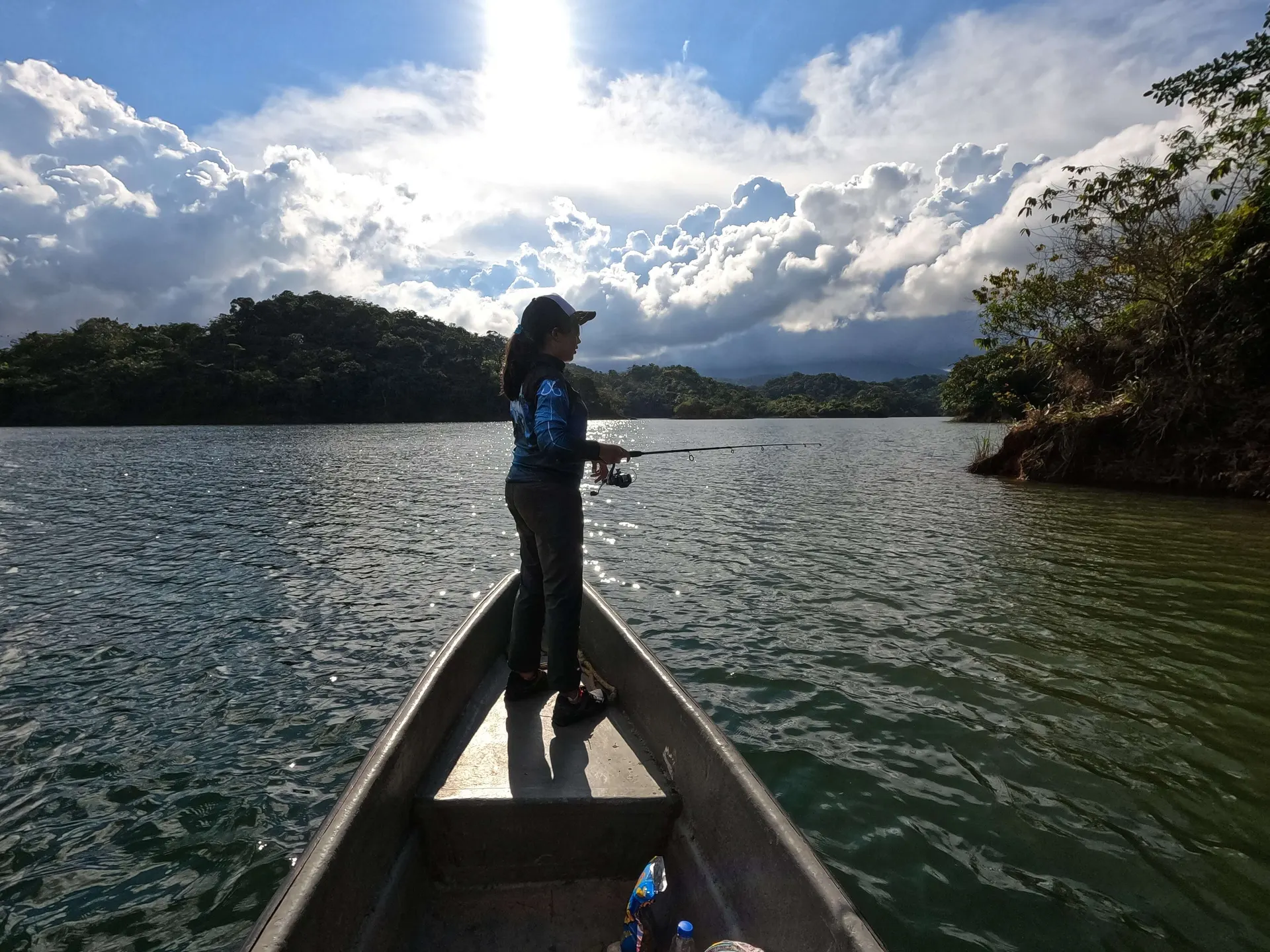 Una pescadora lanza desde la proa de un bote a contraluz, con el sol entre las nubes y las montañas del embalse al fondo.