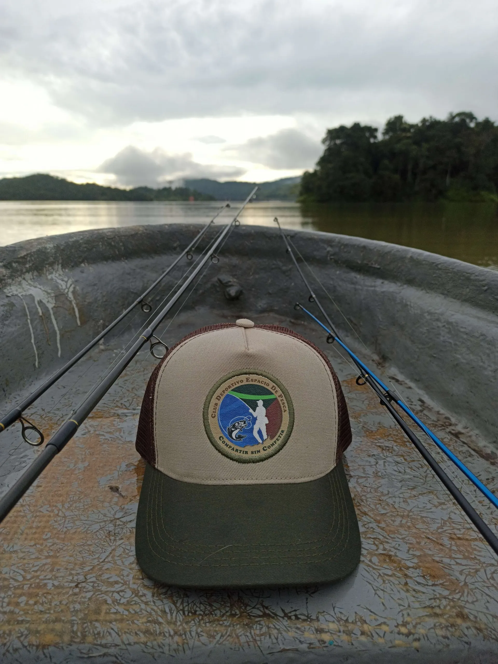 Una gorra del Club Deportivo Espacio de Pesca apoyada en el piso de un bote, con las cañas listas y el embalse al amanecer detrás.