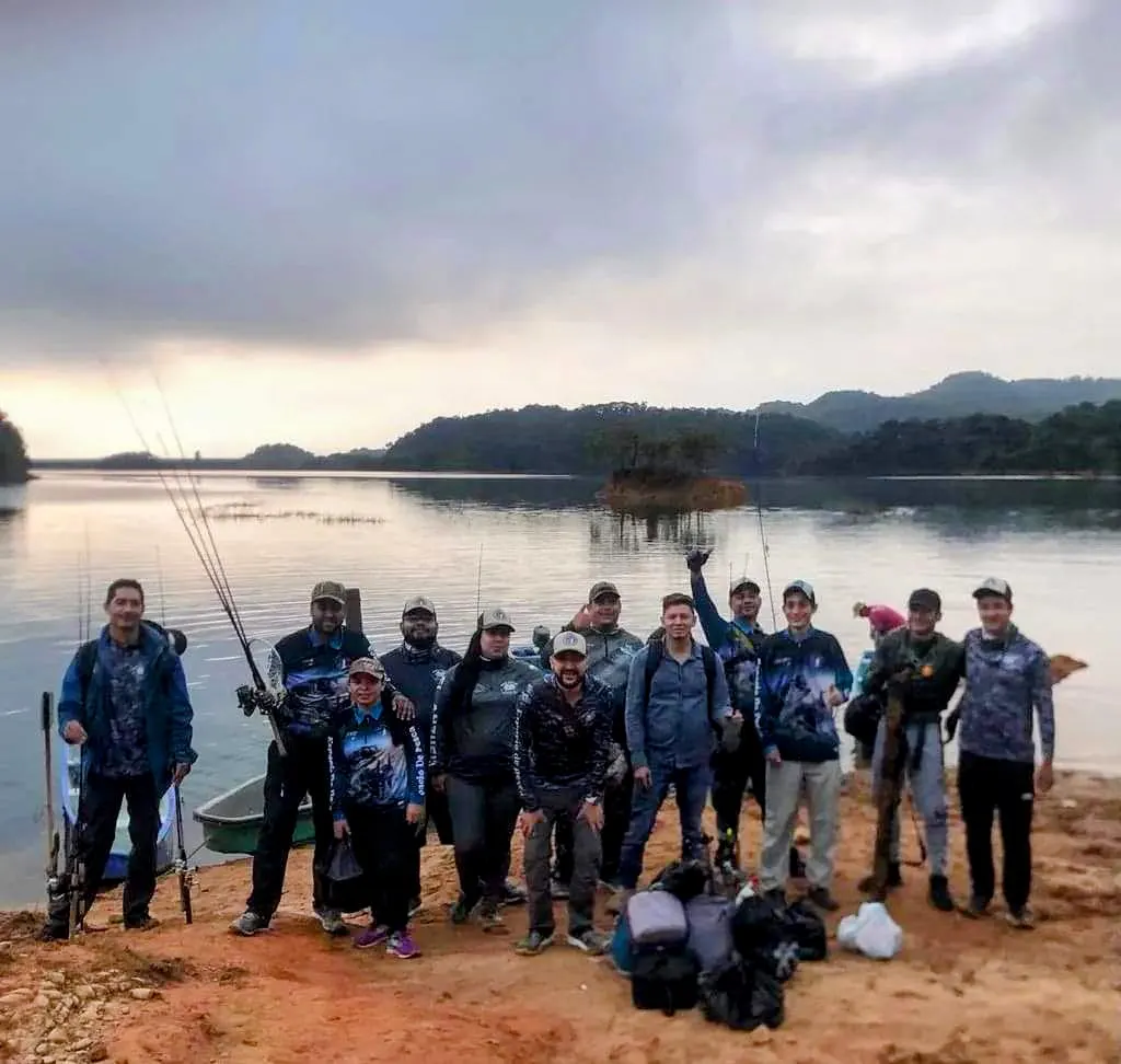 El grupo del club de pesca posa en la orilla del embalse al amanecer, con las cañas y los equipos, bajo un cielo nublado.