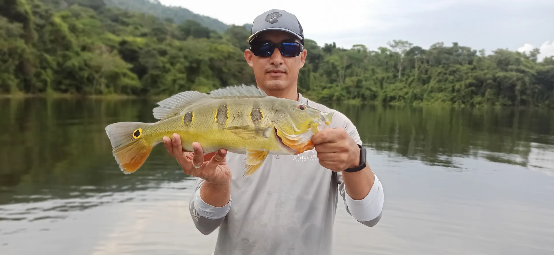 Un pescador con gorra y gafas de sol sostiene con las dos manos un pavón de manchas negras y amarillas, en un embalse rodeado de selva.