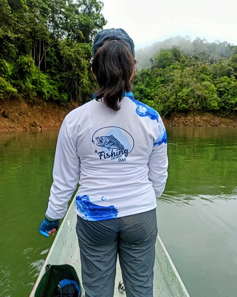 Una joven con camiseta de A Fishing Day sostiene su caña en la proa del bote, con el embalse en calma de fondo.