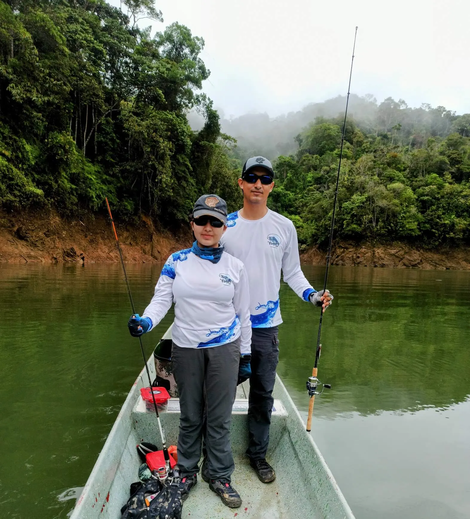 Dos pescadores con camisetas de A Fishing Day posan de pie en un bote con sus cañas, en un embalse rodeado de selva y neblina.