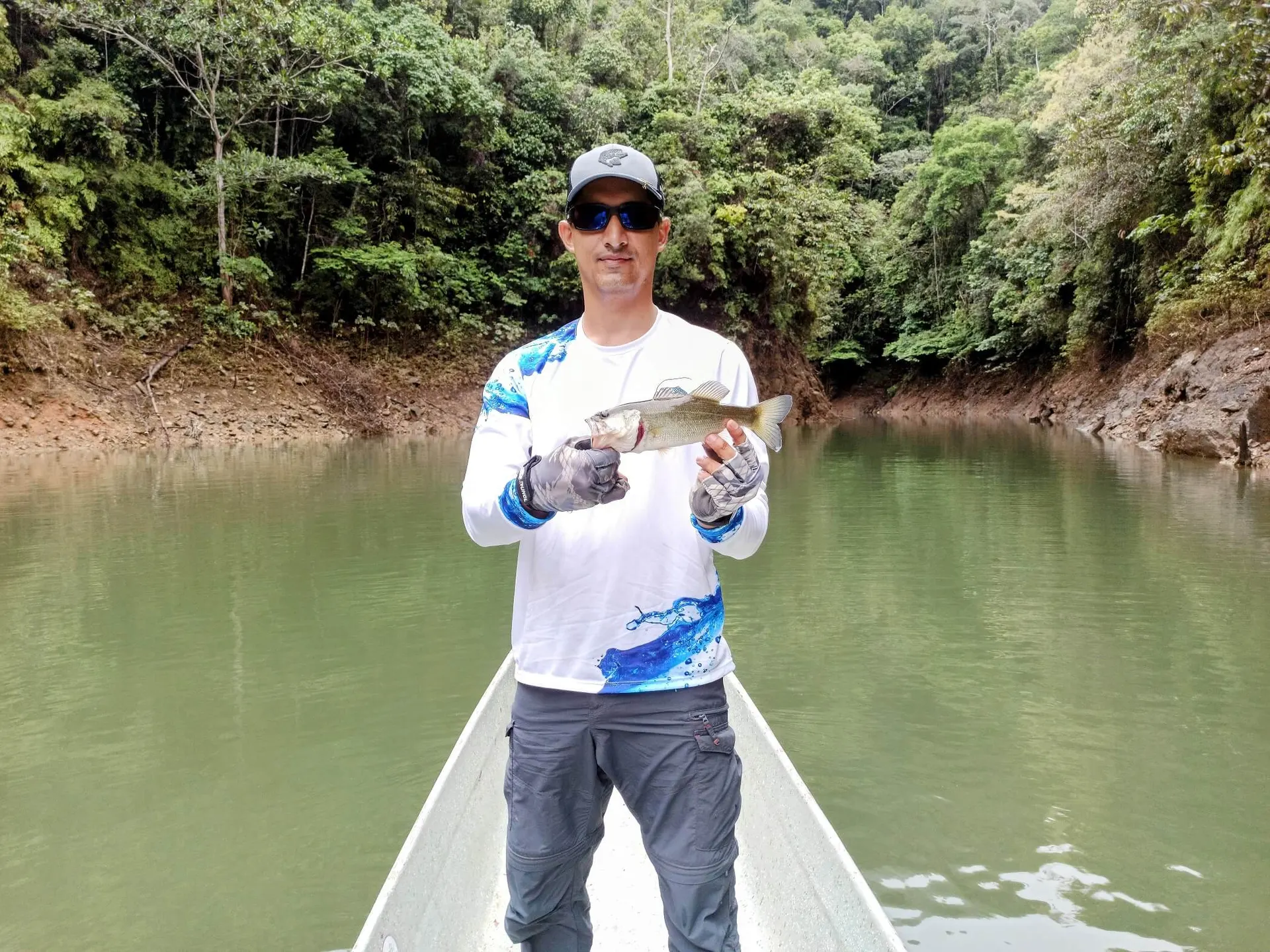 Un black bass es levantado del agua verde del embalse, mostrando su cuerpo oscuro moteado.