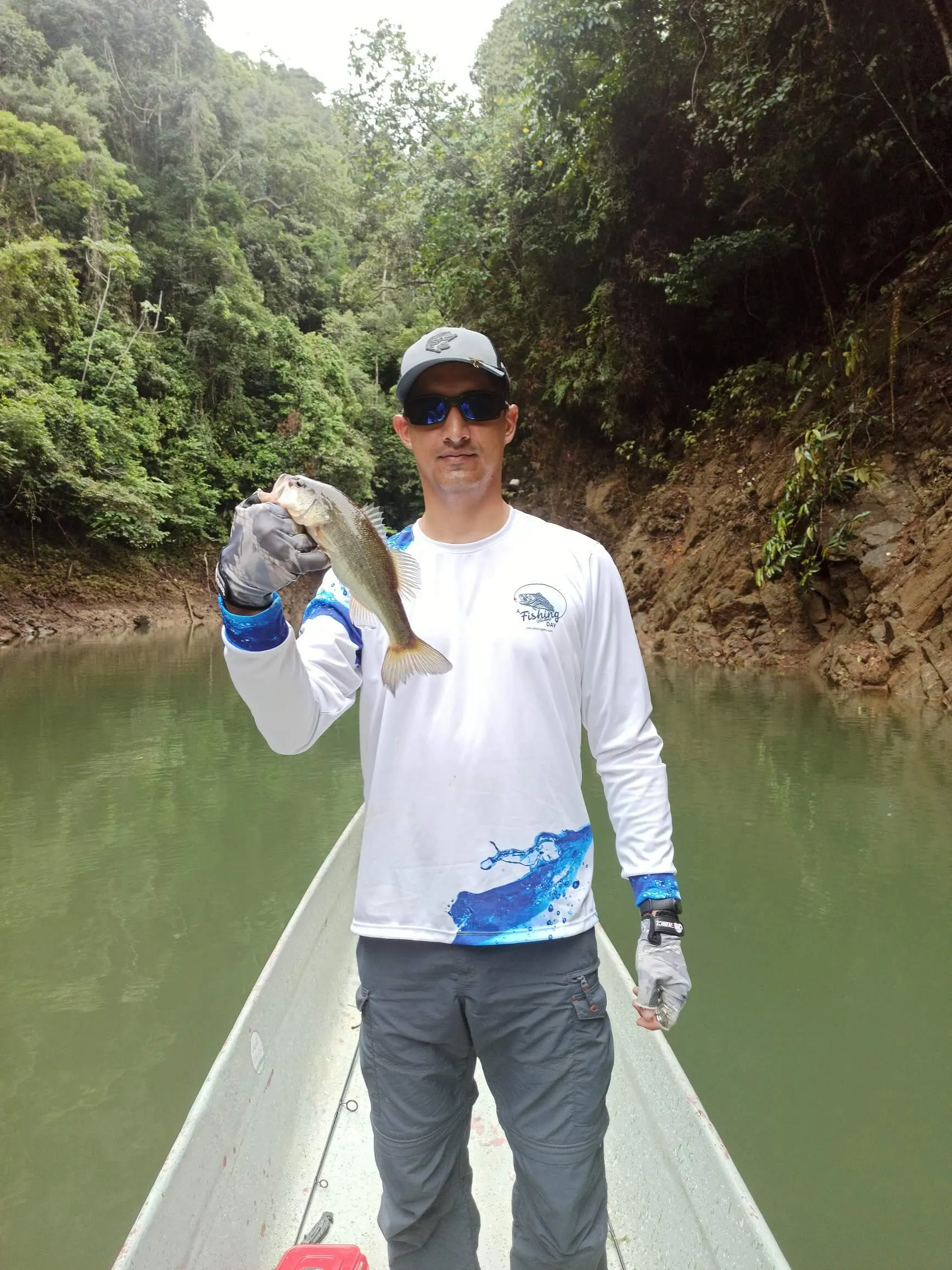 Un pescador con camiseta de A Fishing Day sostiene un black bass recién capturado en la proa del bote.