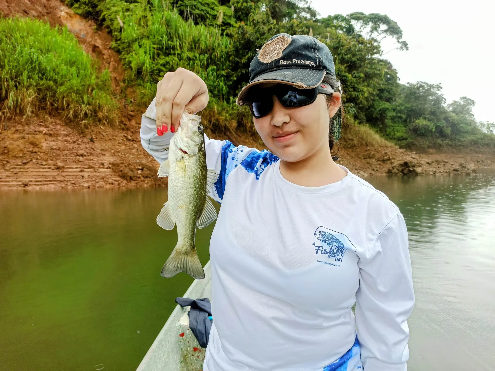 Un pescador sostiene un black bass mediano junto a la orilla del embalse rodeado de vegetación.