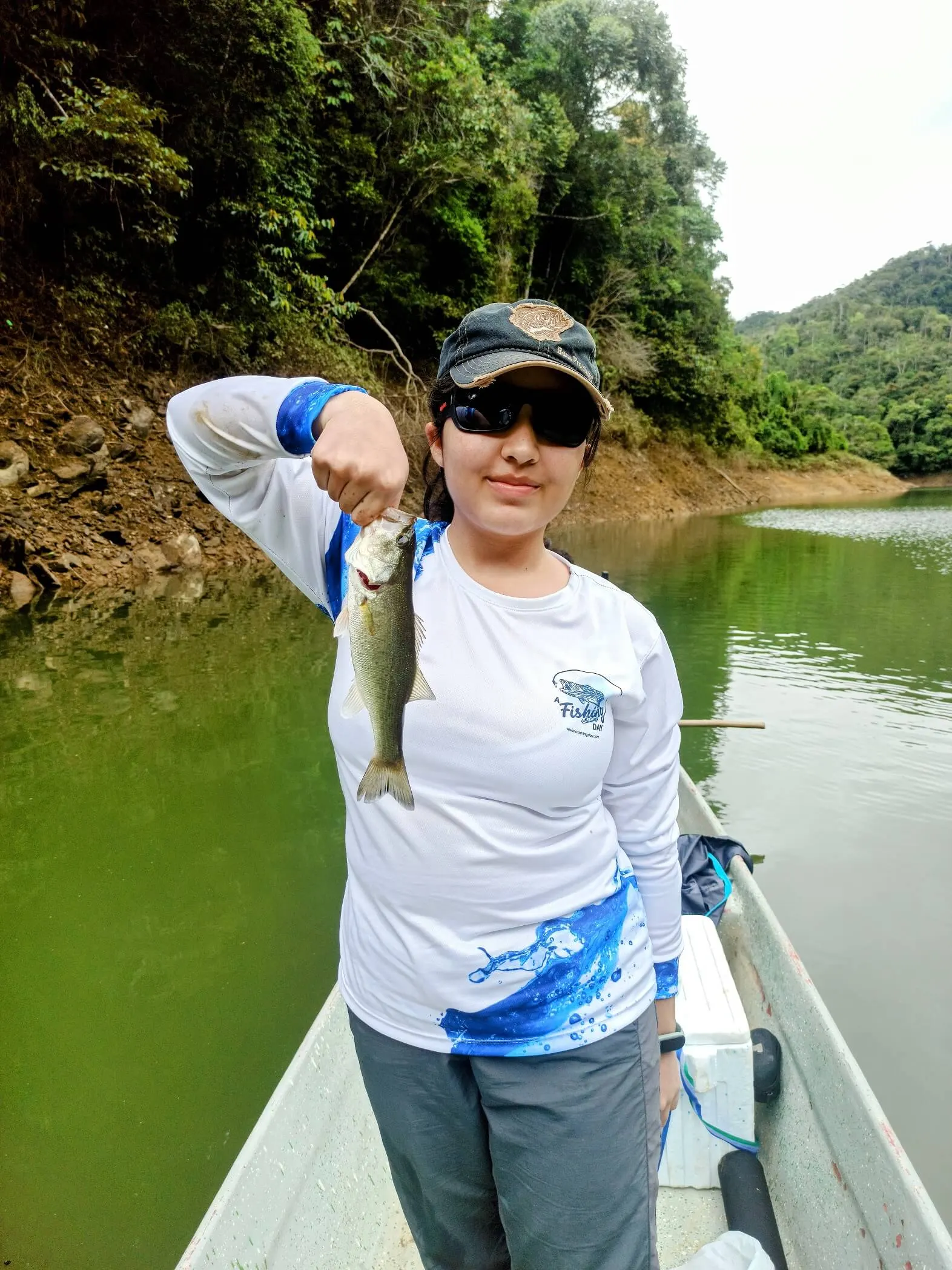 Una joven pescadora levanta un black bass con las dos manos, sonriendo frente al agua tranquila.
