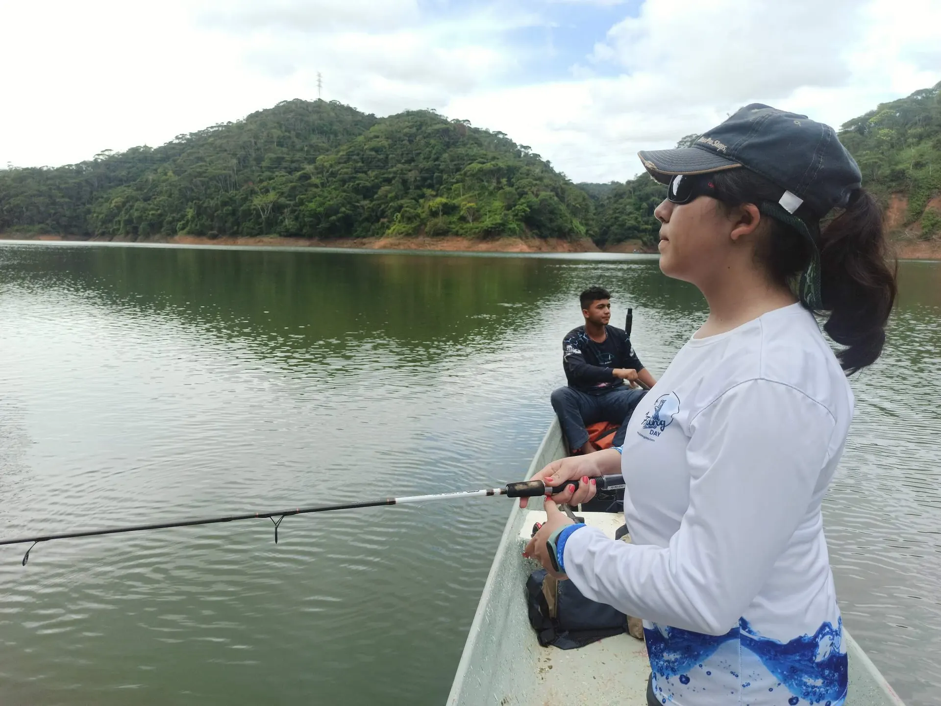 Un pescador sostiene una mojarra plateada capturada en Punchina, con el embalse verde de fondo.