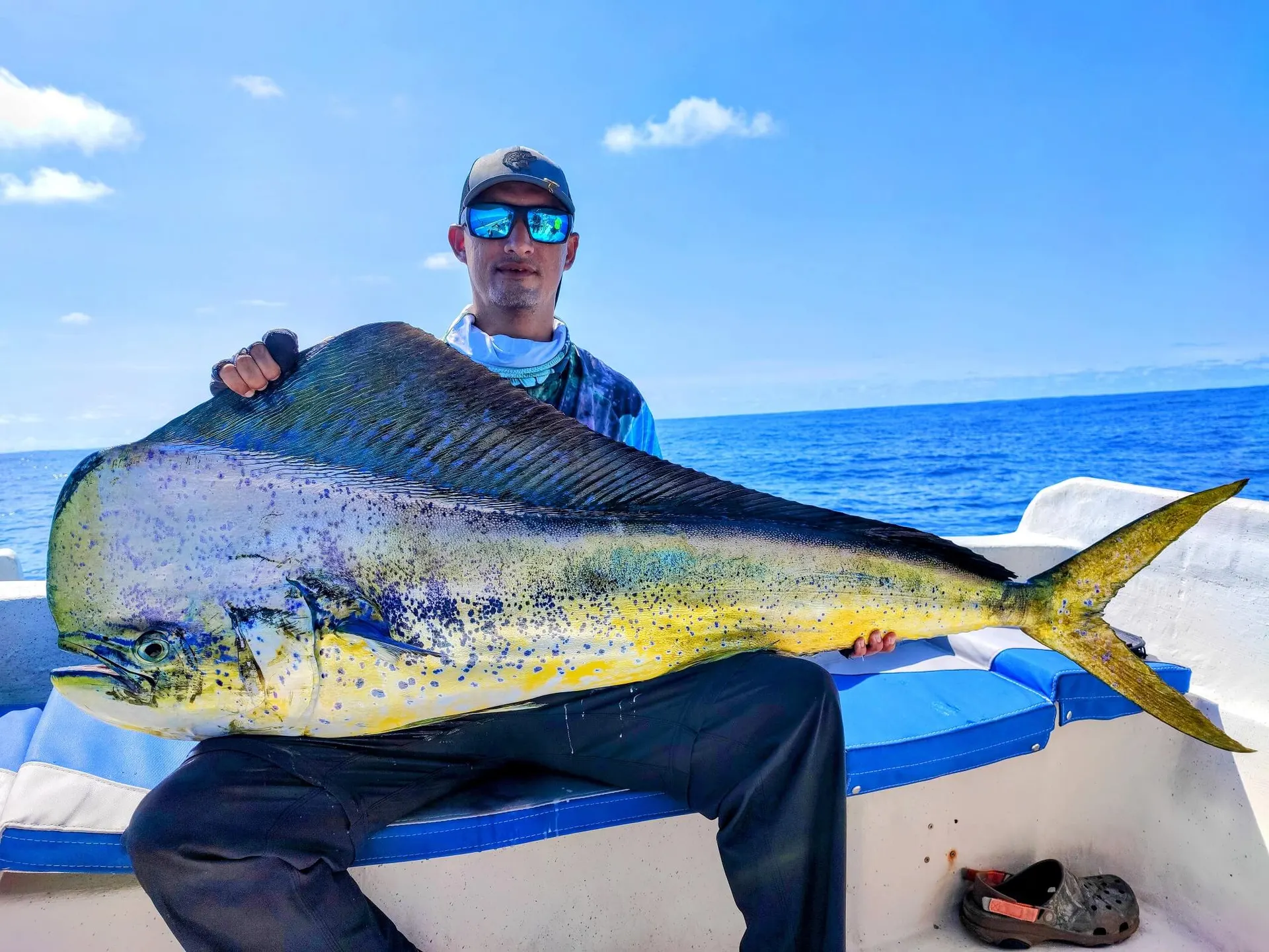Un pescador sentado en un bote sostiene un dorado grande de colores verde, azul y amarillo, en mar abierto bajo un cielo despejado.