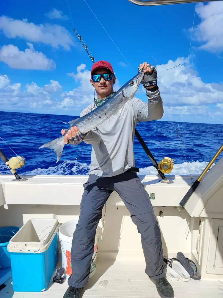 Un pescador sostiene con las dos manos un pez plateado y alargado en la popa de una lancha, en mar abierto de un azul intenso.