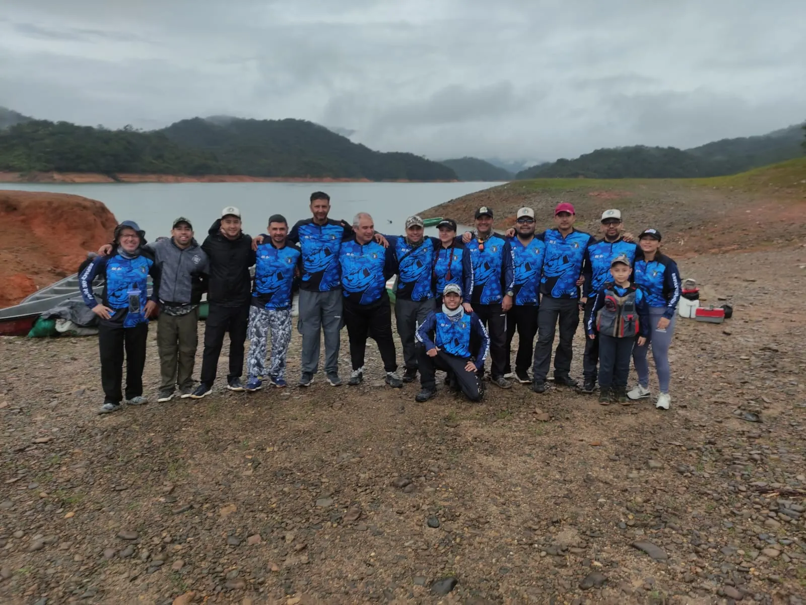 Grupo grande de pescadores con camisetas azules del club posa junto a la orilla del embalse en un día nublado, con las lanchas detrás.