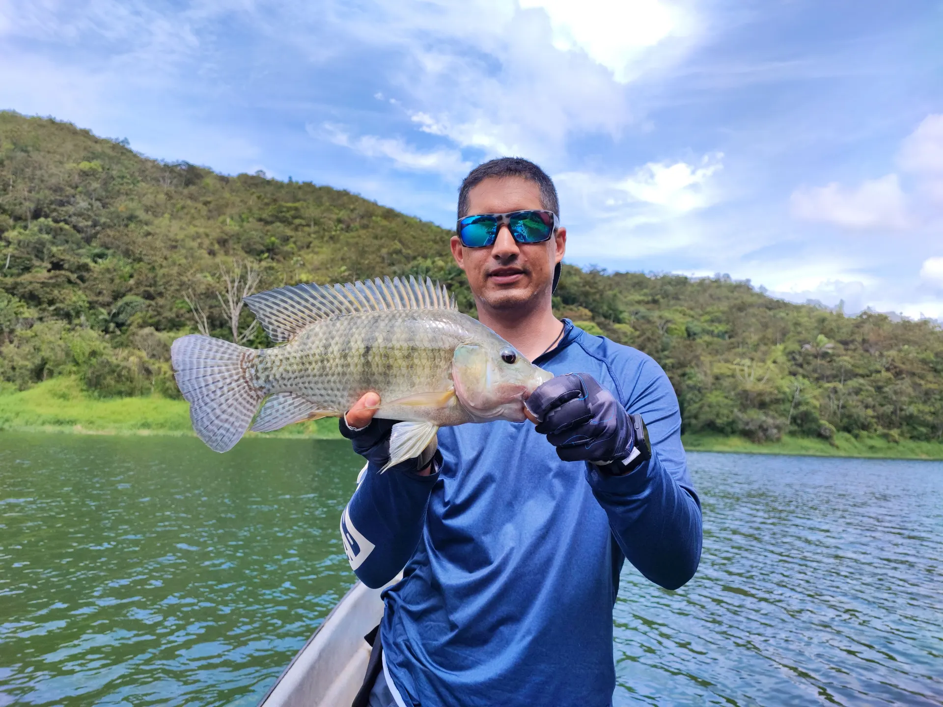 Un pescador con camiseta azul y gafas de sol sostiene una mojarra grande desde un bote, con montañas verdes al fondo.