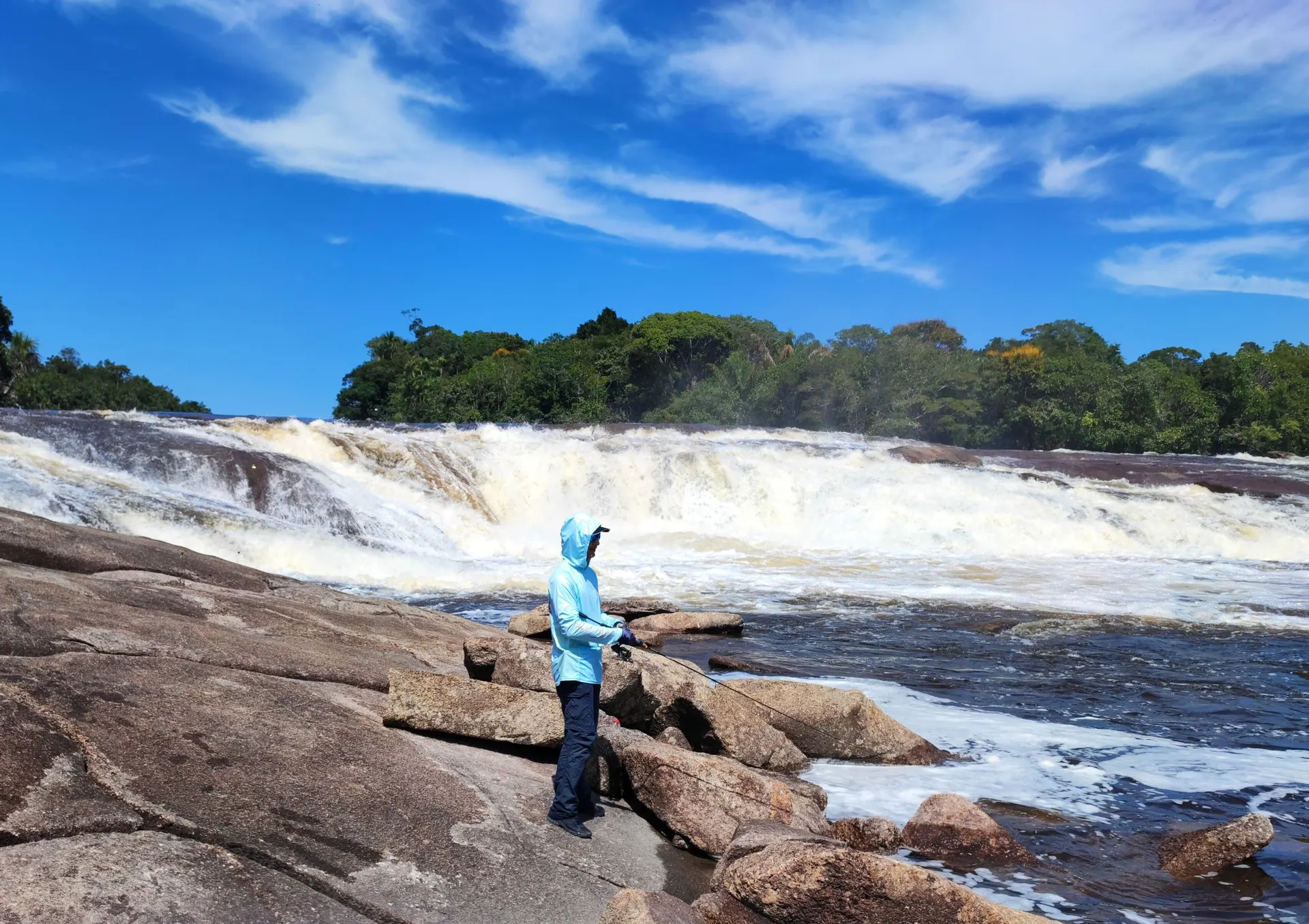 Un pescador con capucha azul clara, de pie sobre rocas de granito, lanza frente a un raudal de aguas blancas bordeado de selva.