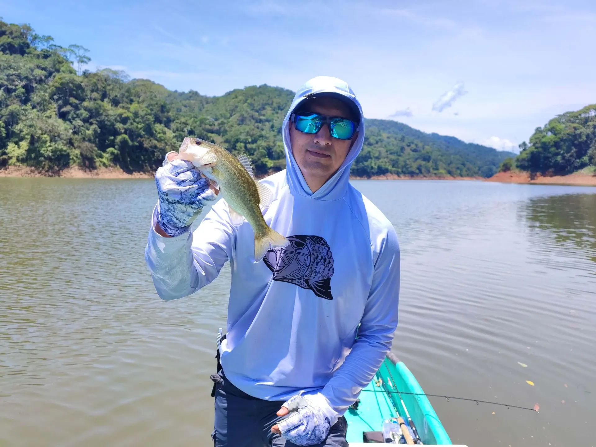 Un pescador con camiseta capucha azul clara y gafas de sol sostiene un black bass pequeño, en un embalse rodeado de montañas selváticas.