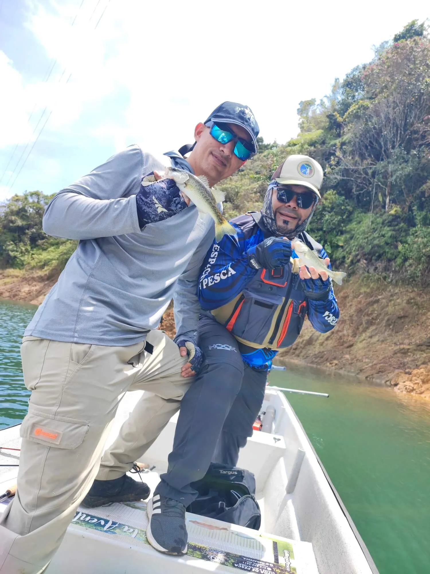Dos pescadores posan en un bote, cada uno mostrando el black bass que acaba de capturar, con la orilla del embalse detrás.