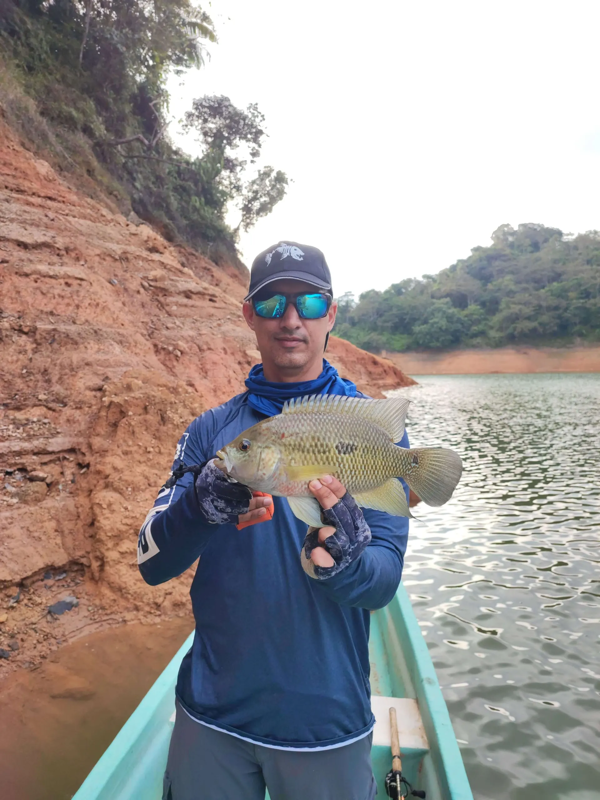Un pescador con camiseta azul y gorra sostiene una mojarra grande desde un bote, con una orilla de tierra roja al fondo.