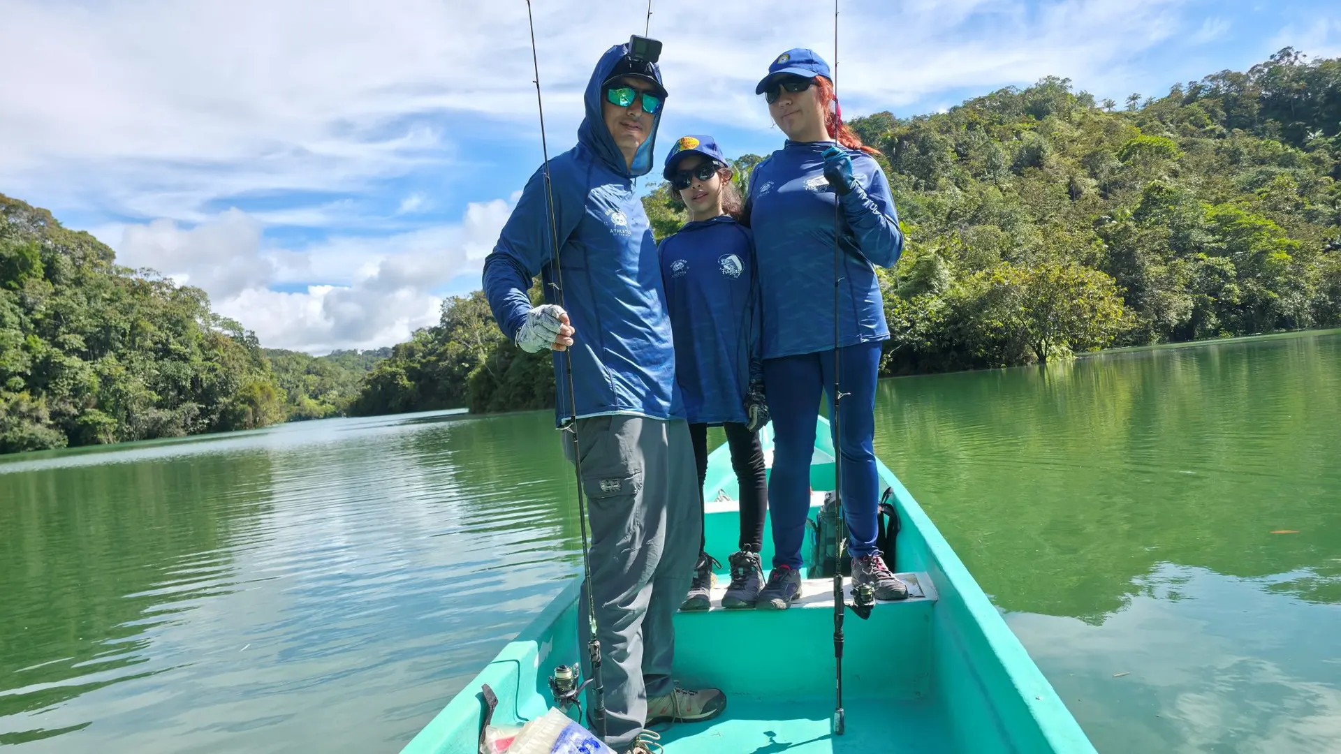 Una familia de tres, con camisetas azules de pesca, posa de pie con sus cañas en la proa de un bote turquesa, en un embalse verde rodeado de selva.